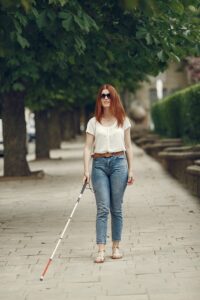 Red-haired woman with sunglasses walking with a white cane on a tree-lined sidewalk